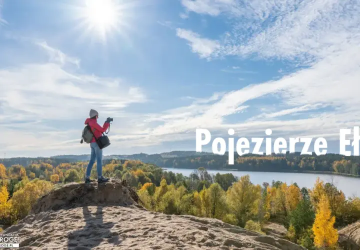 Jesień nad jeziorem. Kobieta z aparatem podziwia krajobraz, szukając ełk atrakcji dla dorosłych. Słońce, chmury i kolorowe drzewa tworzą malowniczy widok.