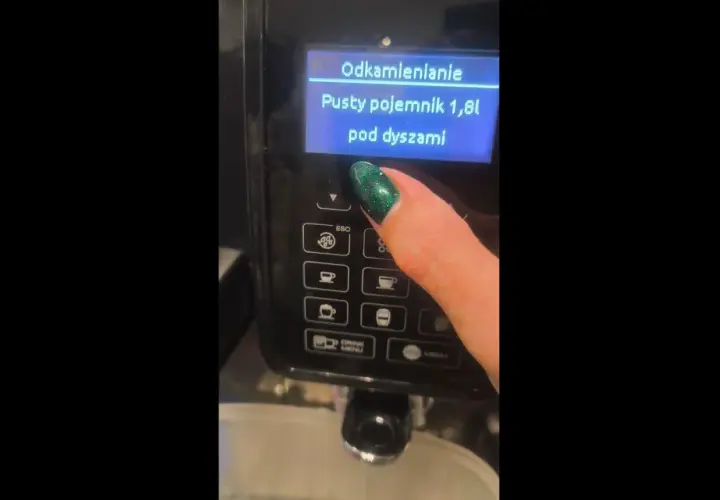 Palec z zielonym lakierem naciska przycisk na ekspresie DeLonghi. Na ekranie wyświetla się komunikat "Odkamienianie. Pusty pojemnik 1,8l pod dyszami".