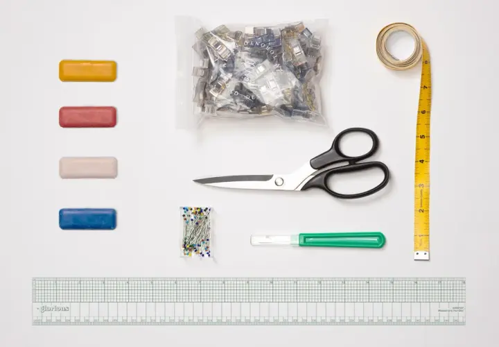 A collection of essential sewing tools, including colorful clips, pins, scissors, a measuring tape, a seam ripper, and a ruler, are laid out on a white surface.
