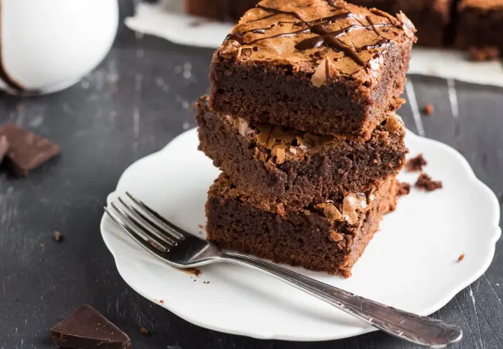 Pyszne ciasto dla odchudzających się: trzy czekoladowe brownie ułożone na talerzyku, obok widelec i kawałki gorzkiej czekolady.