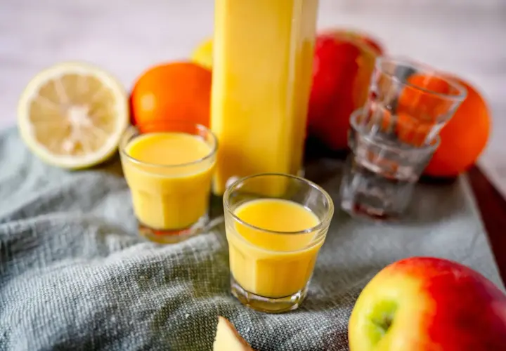 Frische Zutaten für Ingwer Shots selber machen: Zitrone, Orangen, Äpfel und ein Glas voller goldgelbem Shot.