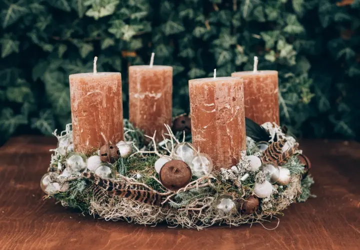 A rustic Advent wreath with four brown candles, moss, feathers, and glass ornaments on a wooden table.