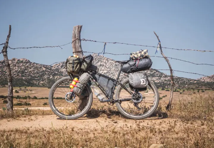 Bicicleta lista para bike packing, cargada con alforjas y bolsas, apoyada en un poste de alambre de espino, con paisaje montañoso al fondo.