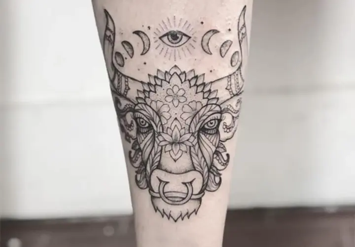 Un detallado tatuaje taurino con un ojo místico y fases lunares sobre la cabeza de un toro adornado con motivos florales y ornamentales.