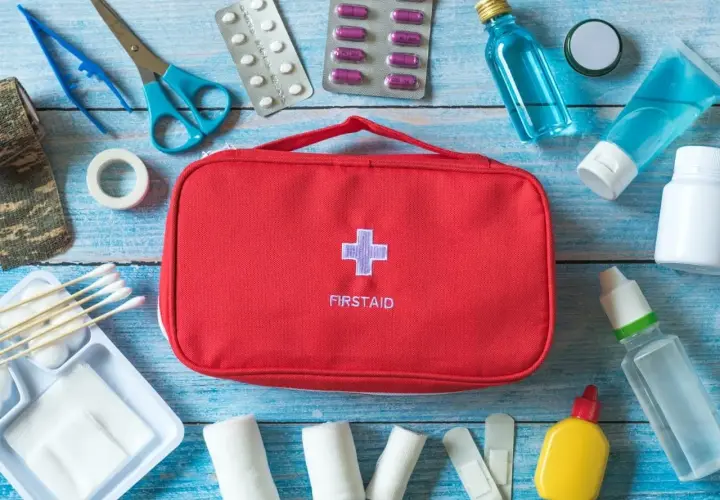 Czerwona apteczka z krzyżem i napisem FIRSTAID otoczona lekami, bandażami, plastrami i akcesoriami medycznymi.