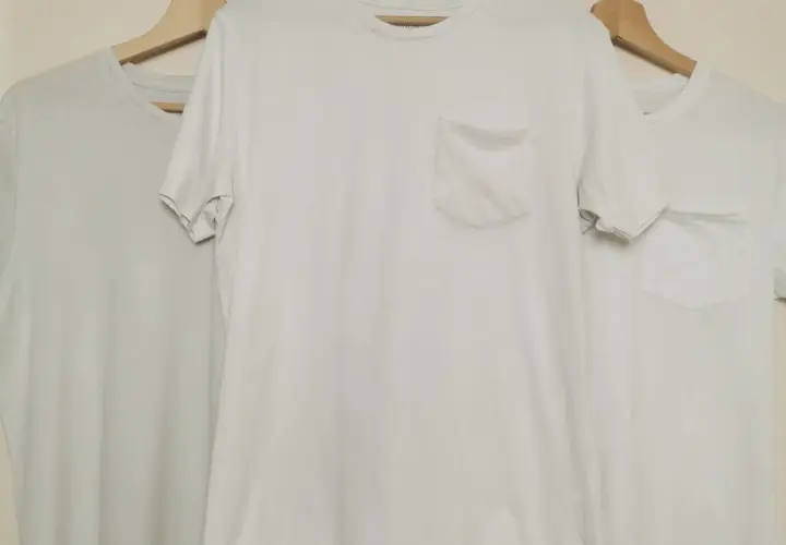 Trois t-shirts blancs sur cintres en bois. Le t-shirt du milieu a une poche poitrine. Idéal pour apprendre comment blanchir un pull blanc.