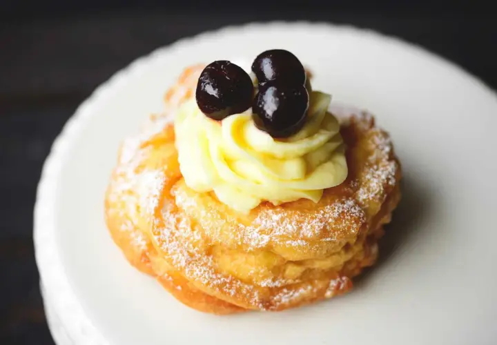 Zeppole fritte gonfie, soffice pasta con crema pasticcera e ciliegie candite, spolverate di zucchero a velo.