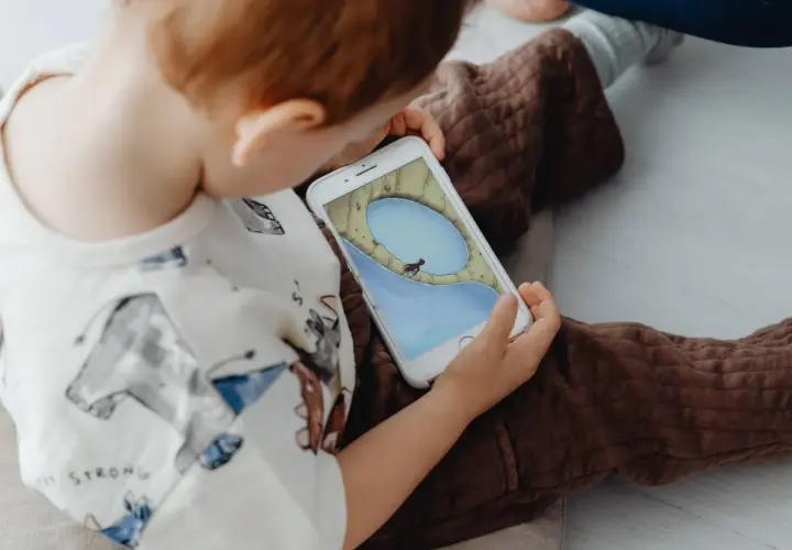 Un niño, absorto en su móvil, parece un niño adicto al móvil. La pantalla muestra un juego con un personaje en un paisaje acuático.