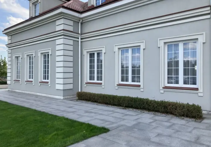 Elegancki budynek z jasnoszarym tynkiem, ozdobiony białymi pilastrami i gzymsami. Okna z białymi ramami i brązowymi parapetami.