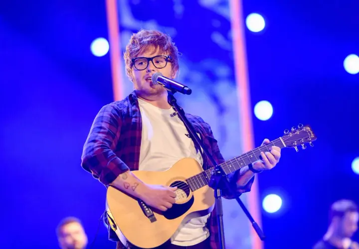 Ed Sheeran w okularach gra na gitarze akustycznej i śpiewa do mikrofonu na niebieskiej scenie.