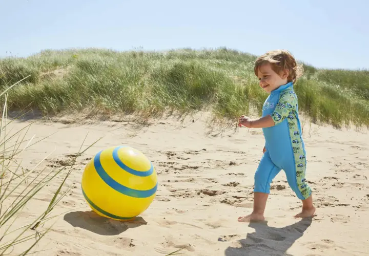 Chłopiec w błękitnym stroju kąpielowym dla niemowląt bawi się na plaży z żółtą piłką.