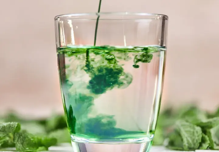 Zielony płyn rozpuszcza się w szklance wody, tworząc wiry. To naturalny chlorofil do picia, otoczony świeżymi liśćmi mięty.