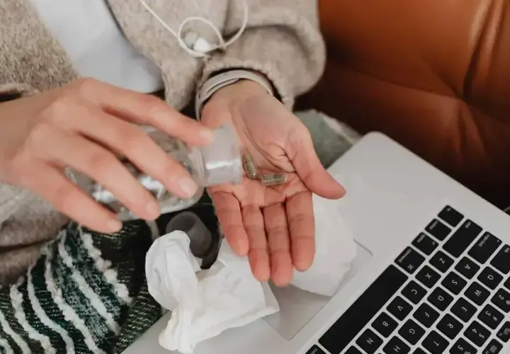 Dłonie z kapsułkami leków, obok laptop i chusteczki. Kto płaci za L4, gdy choroba dopada w domu?