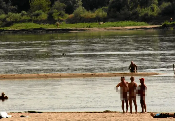 Ludzie opalają się i kąpią na plaży nudystów. Woda jest spokojna, a na brzegu widać kilka osób.