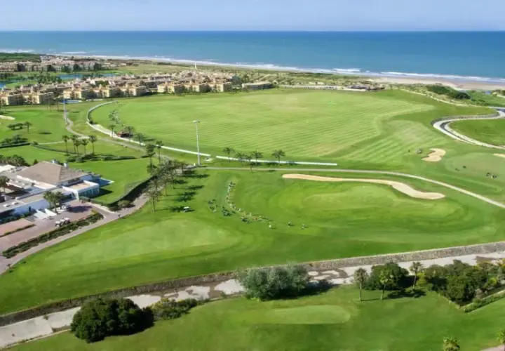 Vista aérea del campo de golf del hotel Barceló Costa Ballena, con el mar azul al fondo y edificios residenciales.