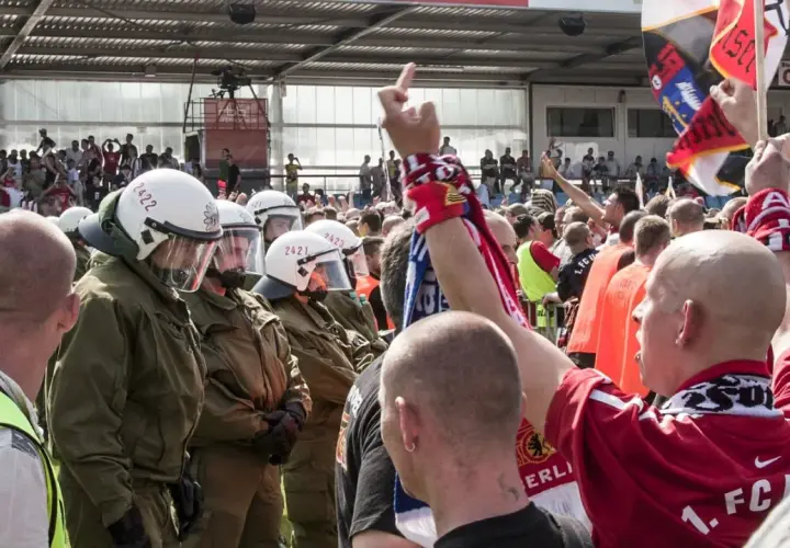 Policja w pełnym rynsztunku stoi naprzeciwko grupy kibiców, wśród których widać ultrasi z flagami i szalikami.