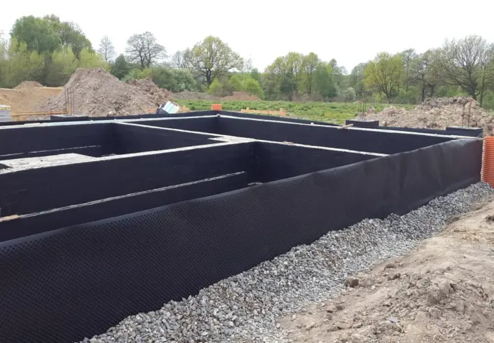 Fundament z czarną izolacją i folią kubełkową. W tle hałdy ziemi i drzewa. To solidna baza, jak izoplast b.