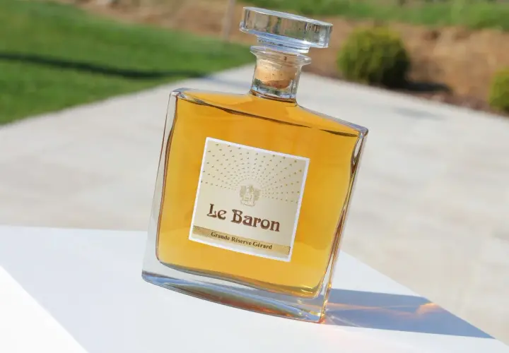 Bouteille de "Le Baron Grande Réserve Gérard", un spiritueux ambré, posée sur un socle blanc, avec un arrière-plan flou évoquant un jardin et un chemin, rappelant l'élégance d'un chateau vaudois.