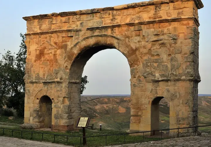 Imponente arco romano de piedra, con dos arcos menores a los lados, se alza sobre un paisaje de colinas verdes.