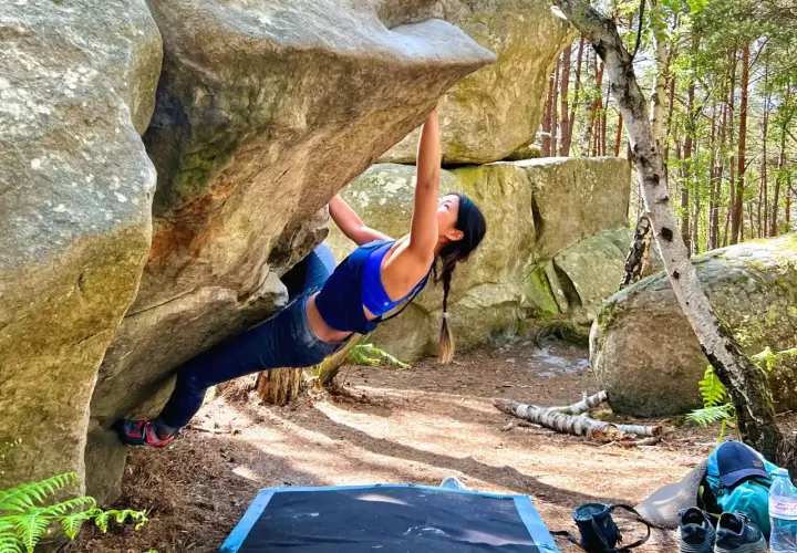 Kobieta wspina się po skale w lesie, doświadczając emocji związanych z boulderingiem w Fontainebleau.