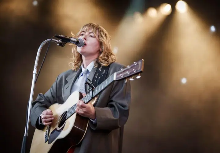 Ellen Krauss konsert. En artist med blont hår, klädd i kavaj och slips, spelar gitarr och sjunger i mikrofonen på en scen med strålkastare.