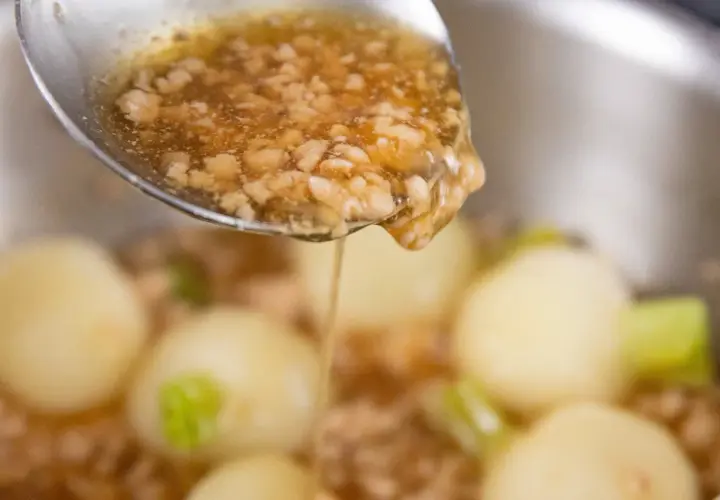 A spoon pours a thick, savory ankake sauce over tender dumplings and vegetables in a pot.