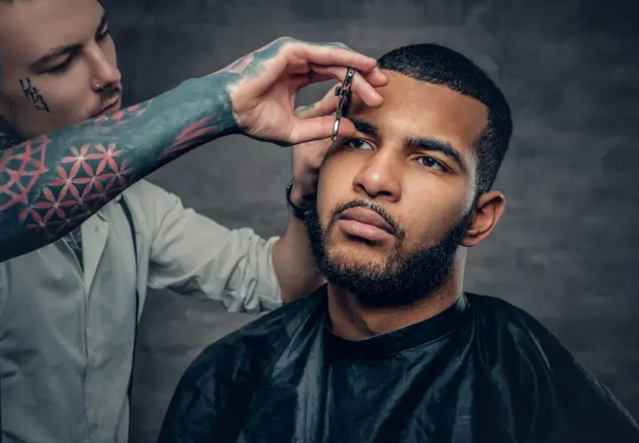 Barbero con tatuajes recorta cejas a hombre con barba, explorando diferentes tipos de buzz cut.
