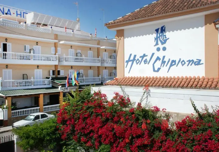 Fachada del Hotel Chipiona con balcones blancos, flores rojas y un coche aparcado.