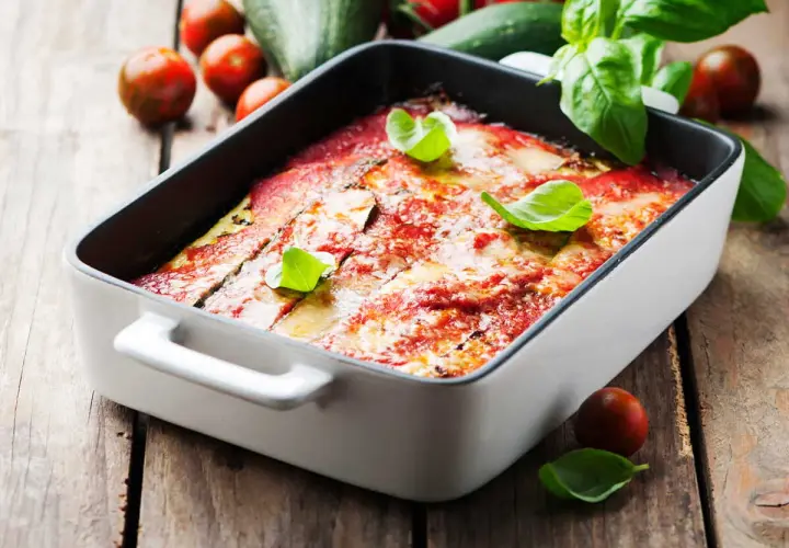 Lasagne courgette gratinée, garnie de basilic frais, sur fond de bois rustique avec des tomates cerises et des courgettes.