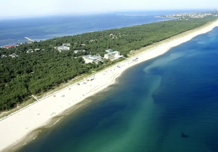 Jurata plaża z lotu ptaka: szeroka, piaszczysta plaża, sosnowy las i budynki hotelowe nad turkusowym morzem.