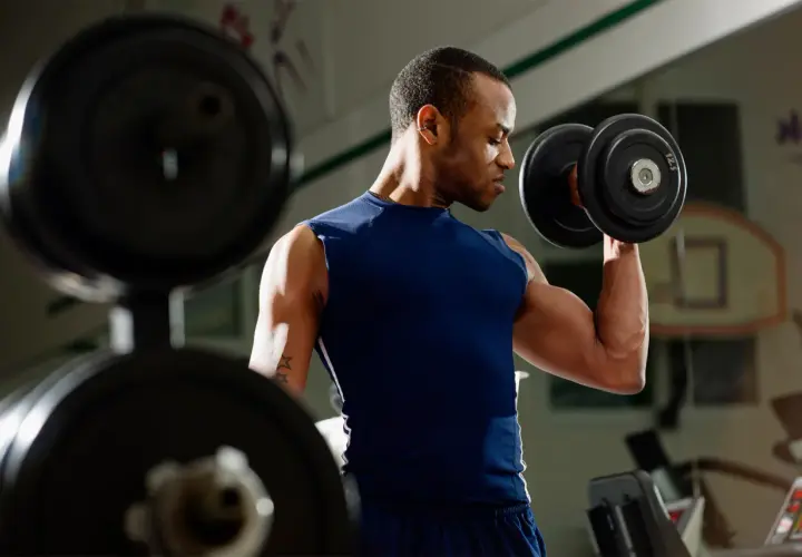 Mężczyzna ćwiczy biceps z hantlem. W tle widać sprzęt do ćwiczeń.