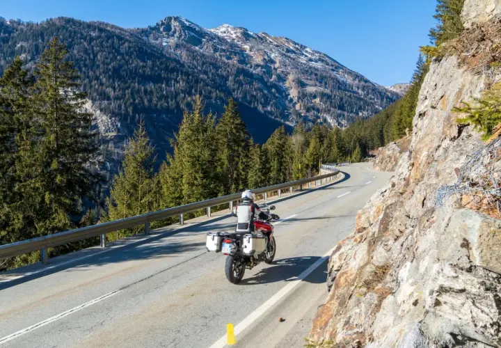 Un motociclista affronta una curva su una strada di montagna, godendosi un perfetto week end in moto tra boschi e cime innevate.