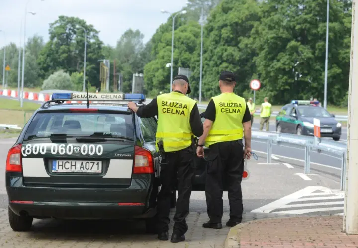 Służba celna czuwa na granicy. Dwóch funkcjonariuszy w kamizelkach odblaskowych stoi przy oznakowanym radiowozie.
