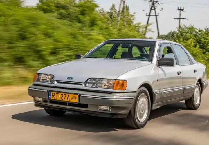 Srebrny Ford Scorpio jedzie po drodze, otoczony zielenią.