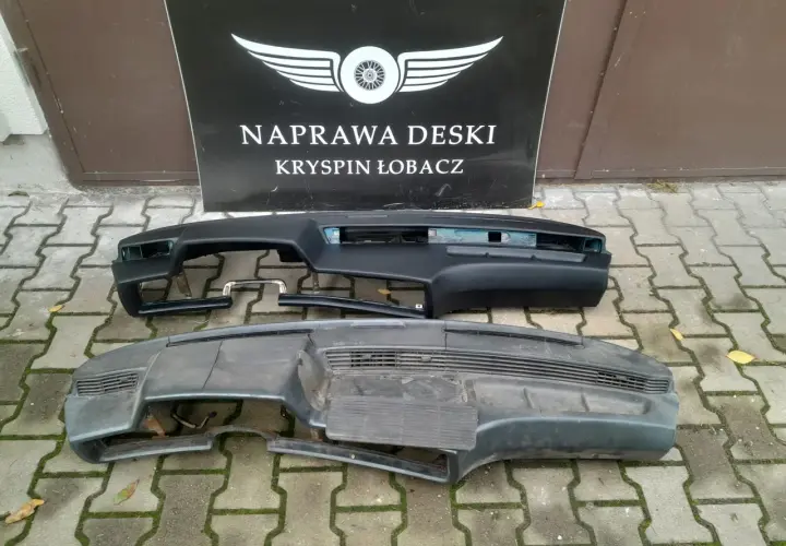 Dwa kokpity samochodowe, jeden czarny, drugi szary, leżą na chodniku przed tablicą z napisem "NAPRAWA DESKI".