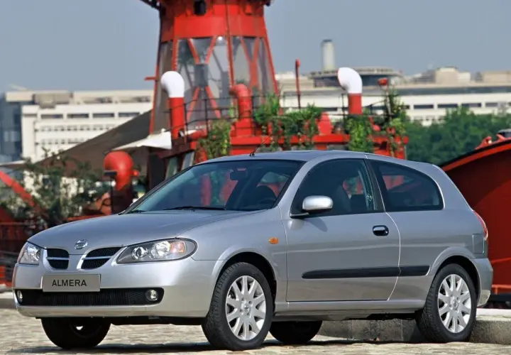 Srebrny Nissan Almera stoi przed czerwoną, industrialną konstrukcją.