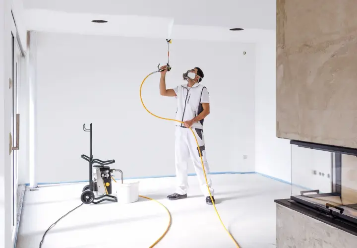 Pittore con maschera protettiva usa una pistola per vernice a spruzzo per dipingere il soffitto di una stanza vuota.
