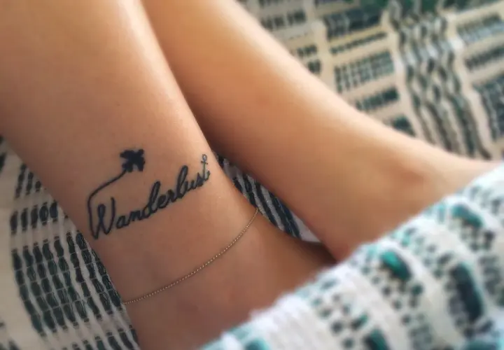 Delikatny tatuaż podróżniczy na nadgarstku: napis "Wanderlust" z samolotem i kotwicą.