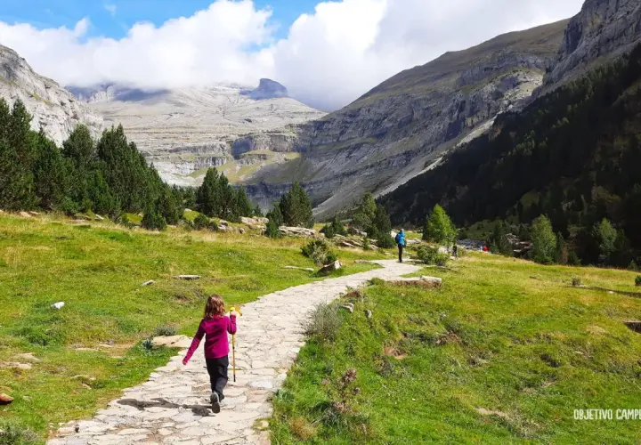 Niños disfrutan de la ruta cola de caballo, un sendero de piedra serpentea por un valle montañoso con pinos y cielos nublados.
