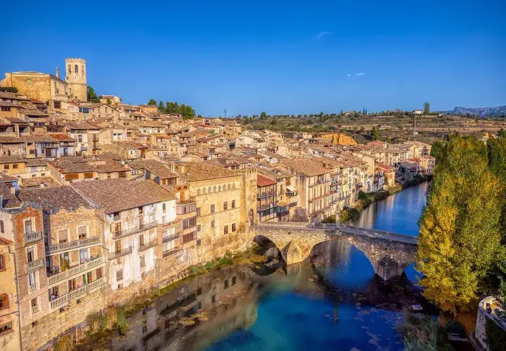 Pueblo con casas de tejas de barro y un puente de piedra sobre un río azul, evocando la toscana española.