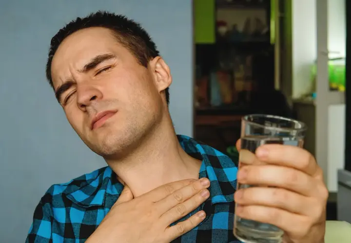 Mężczyzna z zamkniętymi oczami trzyma szklankę wody i dotyka gardła.