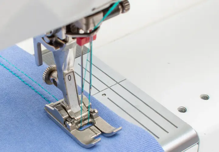 Close-up of a sewing machine needle plate and foot, demonstrating how to use a double needle on blue fabric with turquoise thread.