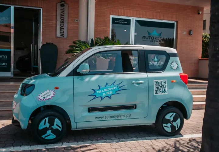 Macchina piccola 4 posti azzurra, con adesivi promozionali "TUA A 99,00 euro AL MESE" e QR code.