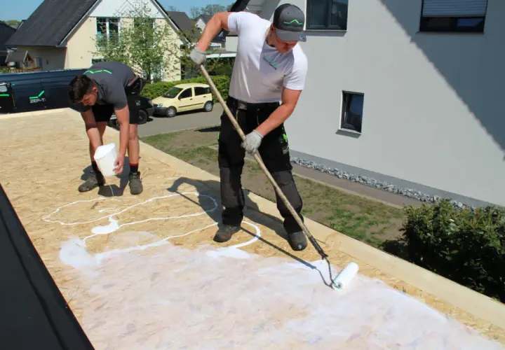 Zwei Handwerker arbeiten auf einem Dach mit OSB-Platten. Einer gießt weiße Flüssigkeit aus einem Eimer, der andere verteilt sie mit einer Rolle.