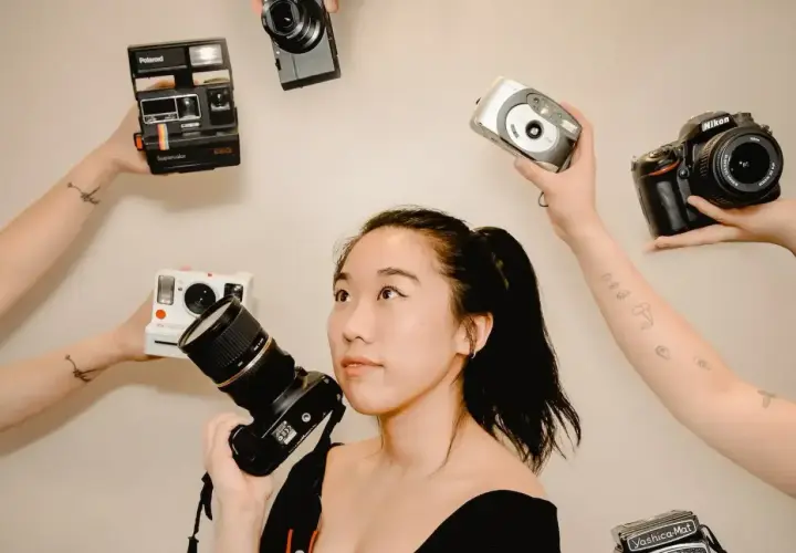 Kobieta z aparatem w ręku, otoczona przez inne aparaty: Polaroid, Nikon, cyfrówki. Idealna do wyboru, jaka lustrzanka na początek.