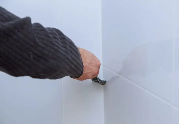 Dłoń w swetrze usuwa silikon z białych płytek za pomocą narzędzia.