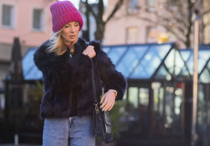 Krótkie futerko stylizacje: blondynka w różowej czapce i czarnym futerku, z jeansami i torebką, na tle miasta.