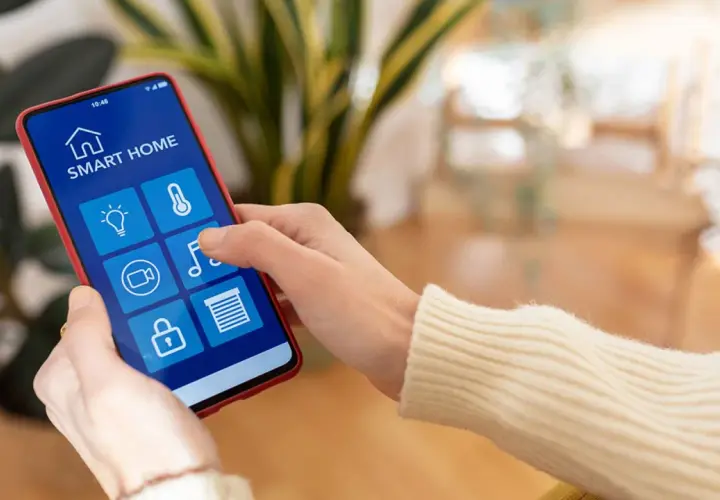 Mano usando un smartphone para controlar productos inteligentes de hogar: luces, temperatura, cámara, música y persianas.