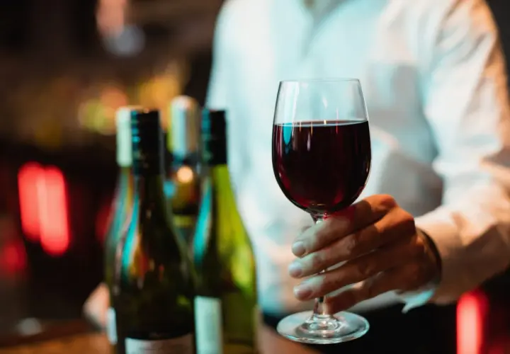 Main d'un homme tenant un verre de vin rouge, prêt pour une dégustation de vin Beaujolais. Des bouteilles floues en arrière-plan.