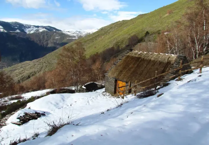 Pequeña cabaña de piedra con tejado de paja, rodeada de nieve y un paisaje montañoso. Un esterri de cardos se alza en la ladera verde.